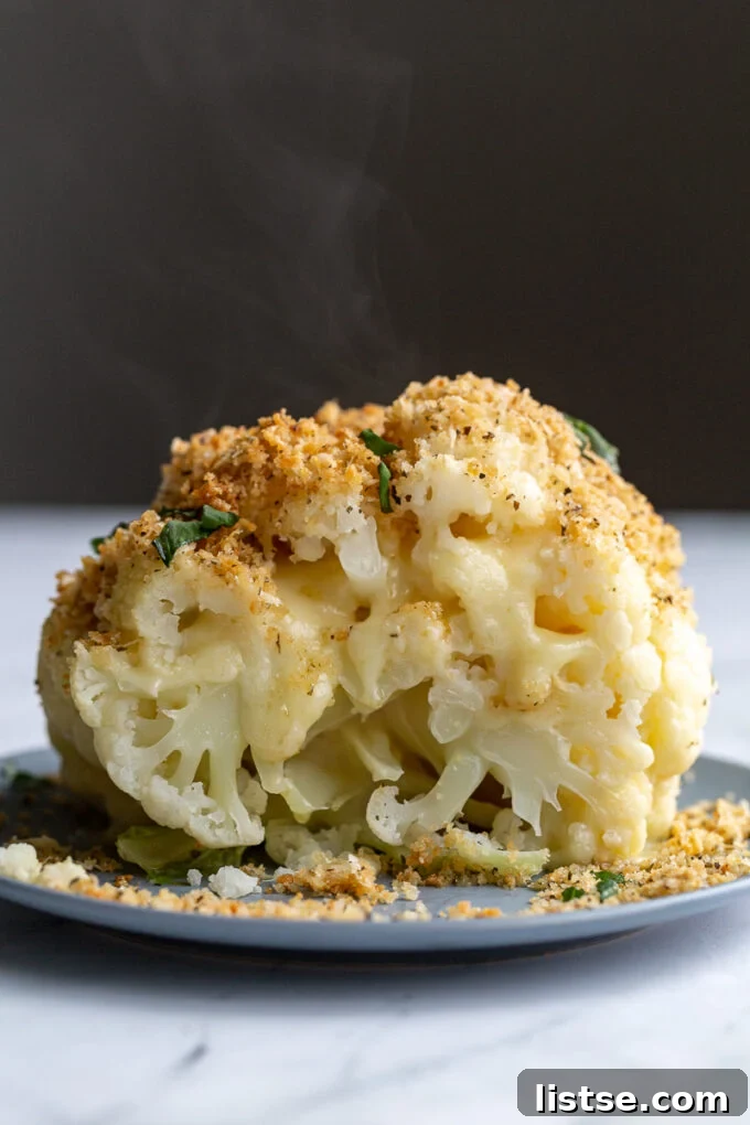 A half-cut whole head of roasted cheesy cauliflower, showcasing creamy melted cheese oozing from the florets, topped with crunchy golden breadcrumbs and fresh basil, presented beautifully in a blue dish.