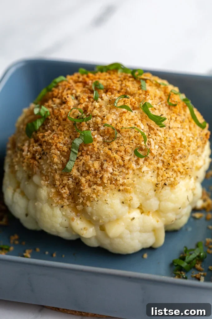 A whole head of cheesy stuffed cauliflower baked to golden perfection in a blue baking dish, with melted cheese visible between the florets, topped with a delicious toasted breadcrumb and basil garnish.