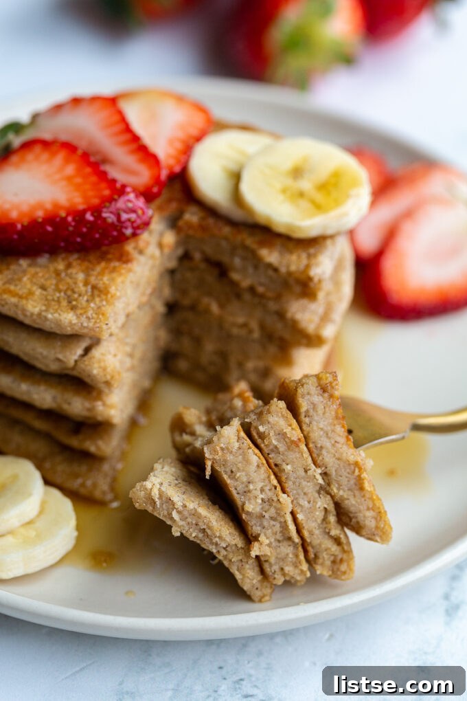 stack of oat flour pancakes on a plate that are topped with banana and strawberry slices and maple syrup. More banana and strawberries on the side of the plate. A bite has been taken out of the pancakes and the stacked bite of pancakes is resting on the plate
