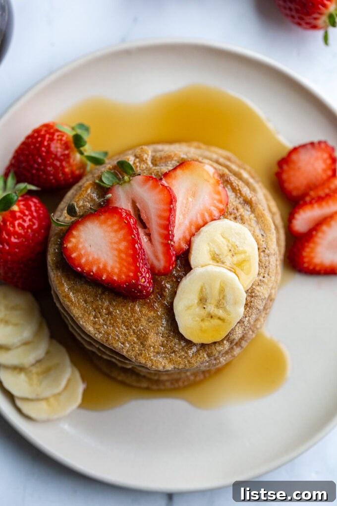 stack of oat flour pancakes on a plate that are topped with banana and strawberry slices and maple syrup. More banana and strawberries on the side of the plate