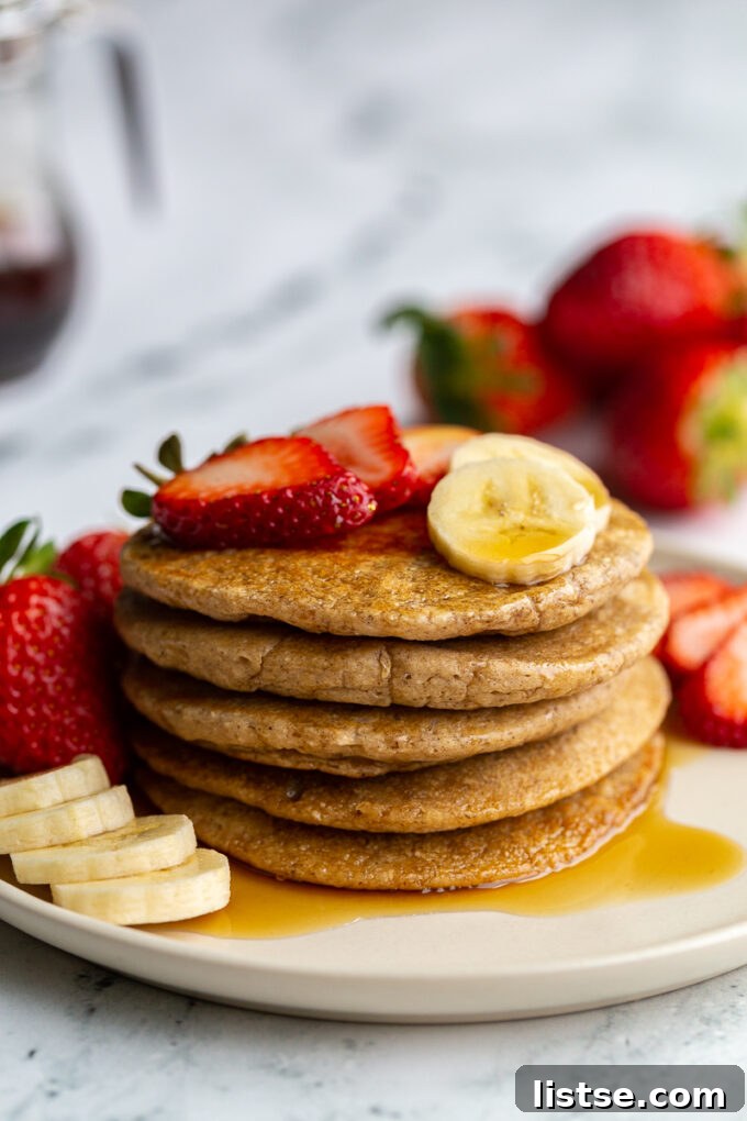stack of oat flour pancakes on a plate that are topped with banana and strawberry slices and maple syrup. More banana and strawberries on the side of the plate