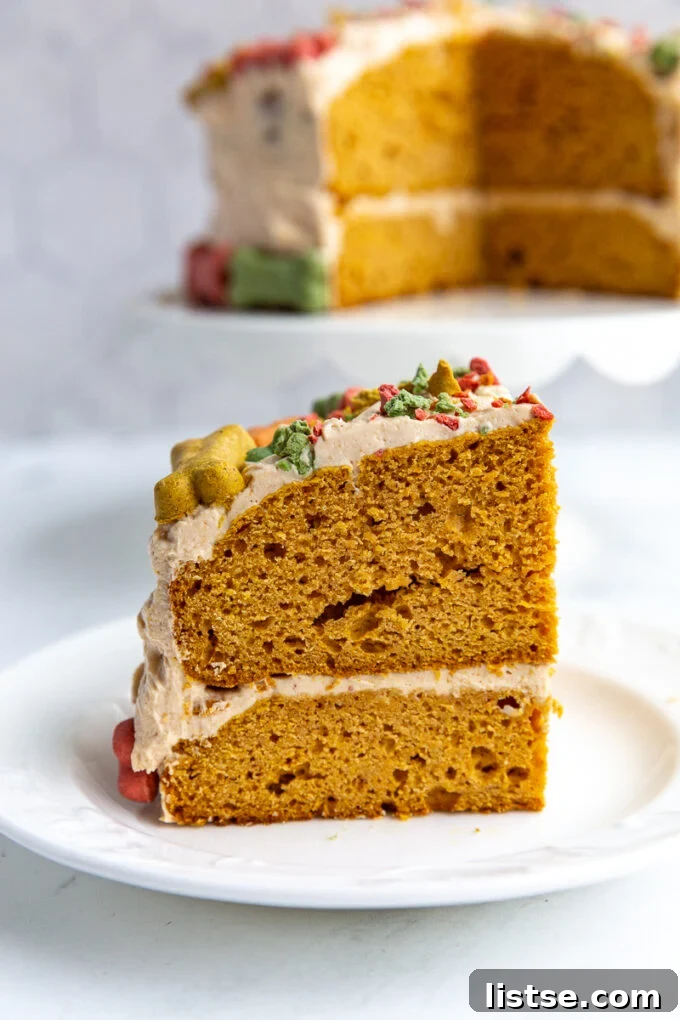 A perfectly portioned slice of dog birthday cake sits on a plate, with the remainder of the festive cake displayed on a cake stand in the background. The cake is adorned with colorful dog bones, hinting at a delightful celebration.