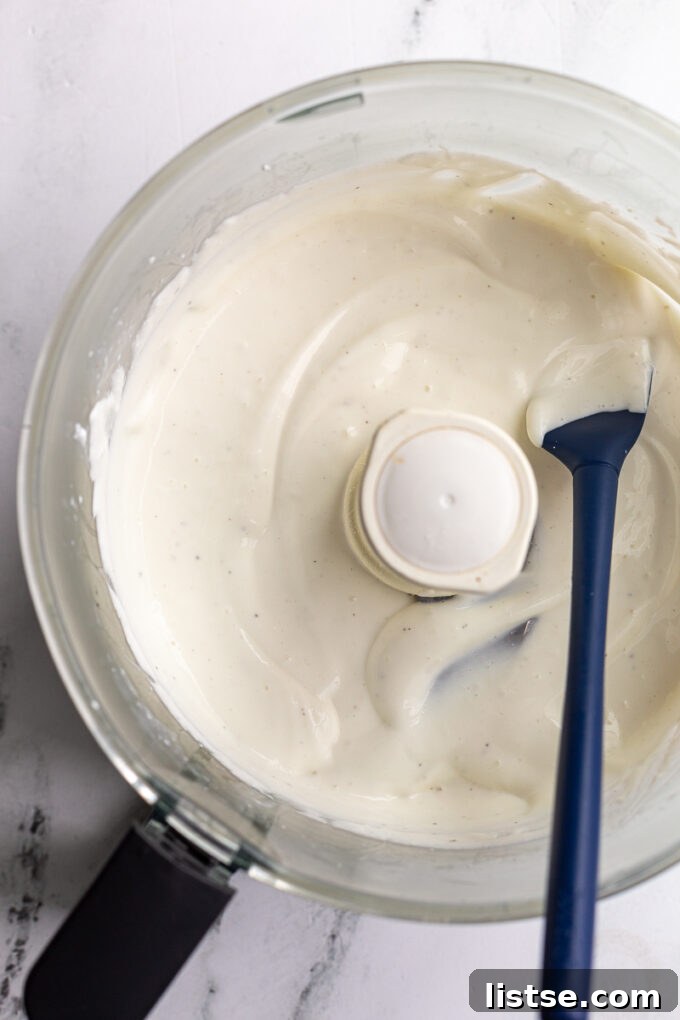 Creamy Cottage Cheese Celery Boats 4 A food processor, with its lid removed, contains creamy whipped cottage cheese. A navy blue spatula rests on the side, ready to assist in the blending process.