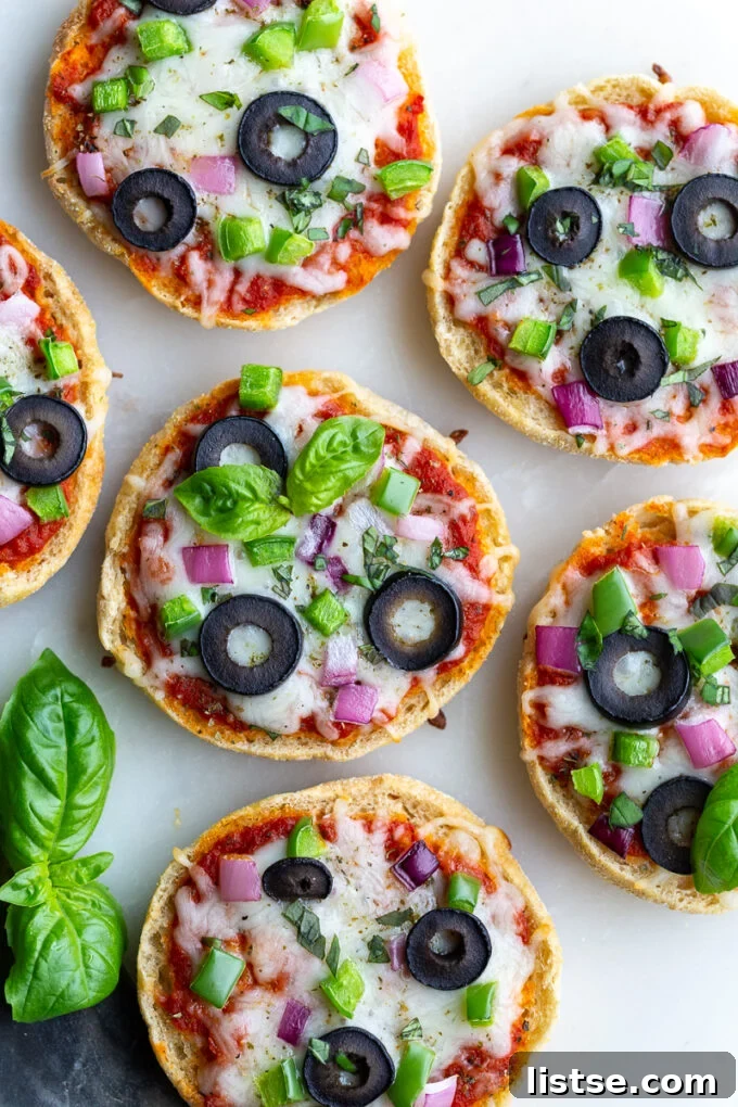 Close-up shot of English Muffin Pizzas, topped with melted cheese, vibrant red onion, crisp green pepper, briny olives, and fresh basil, resting on a pristine white board.