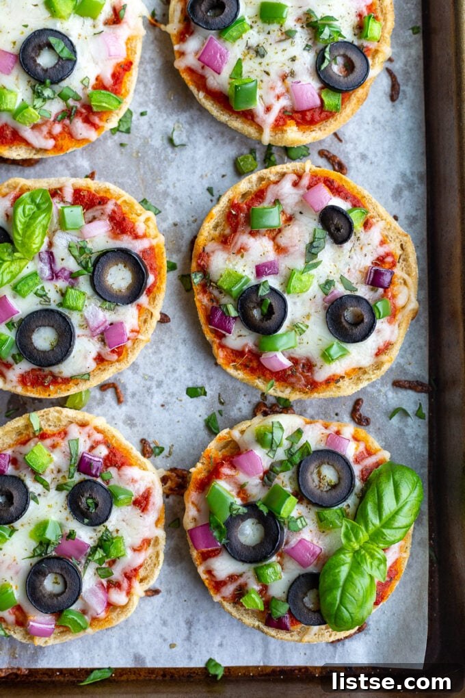 Close-up shot of English Muffin Pizzas, generously topped with cheese, red onion, green pepper, olives, and fresh basil, arranged neatly on a parchment-lined baking sheet, ready for the oven.