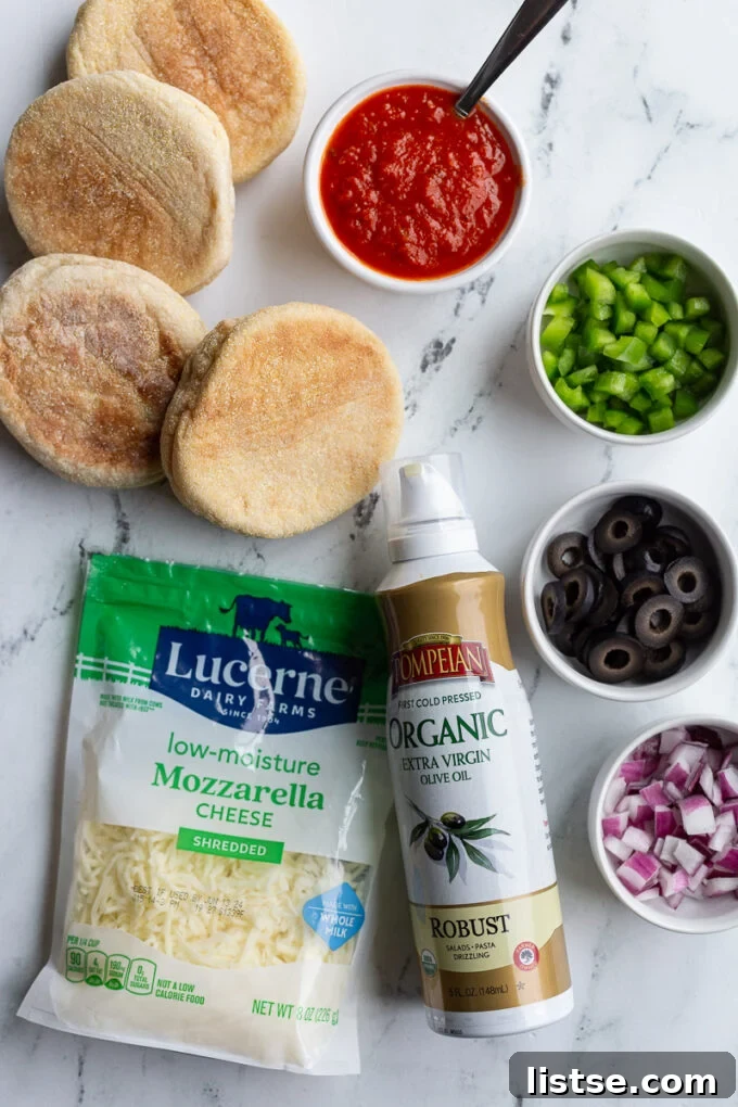 Ingredients laid out for English Muffin Pizzas: halved English muffins, olive oil cooking spray, shredded mozzarella cheese, finely diced red onions, black olives, and bell pepper, alongside a bowl of rich pizza sauce.