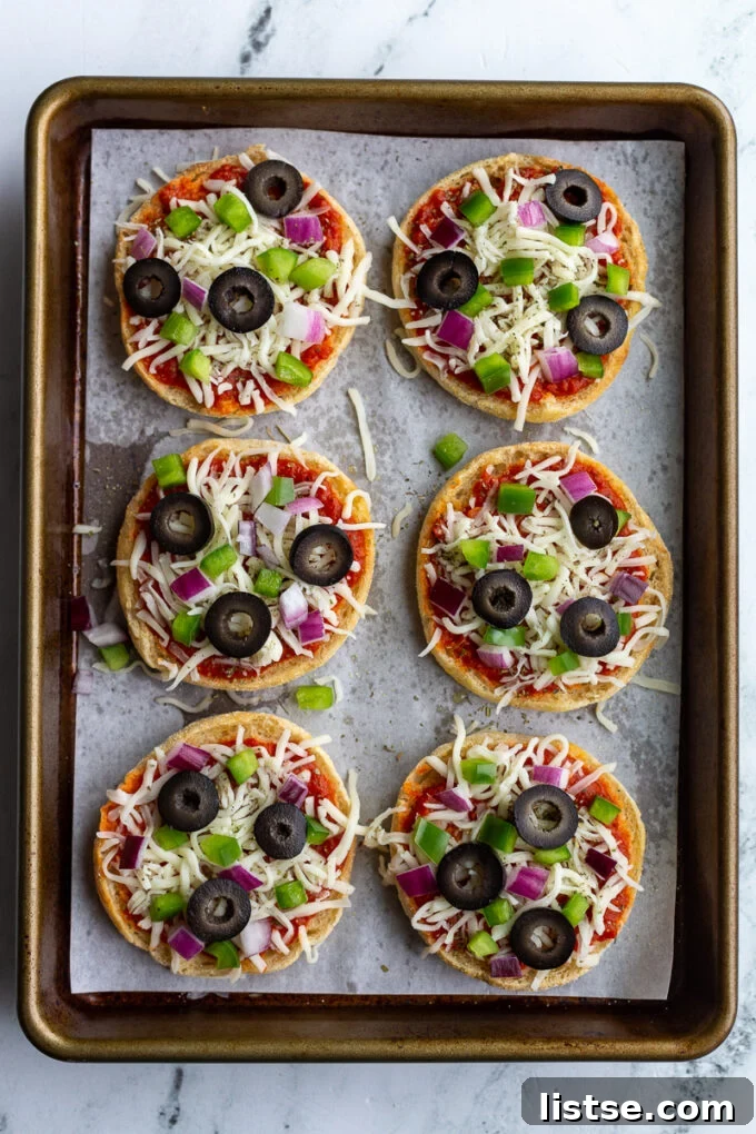 Unbaked English Muffin Pizzas, topped with cheese, red onion, green pepper, olives, and basil, arranged on a parchment-lined baking sheet before being placed in the oven.