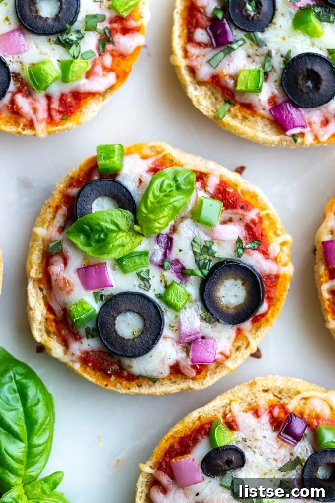 Close-up shot of English Muffin Pizzas, topped with cheese, red onion, green pepper, olives, and basil, sitting on a clean white board after being baked to perfection.