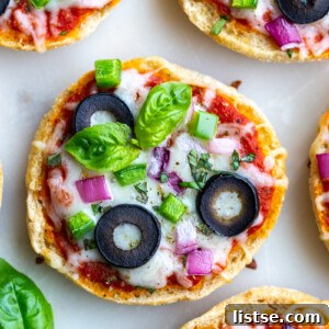 Close-up shot of English Muffin Pizzas, topped with cheese, red onion, green pepper, olives, and basil, sitting on a white board.