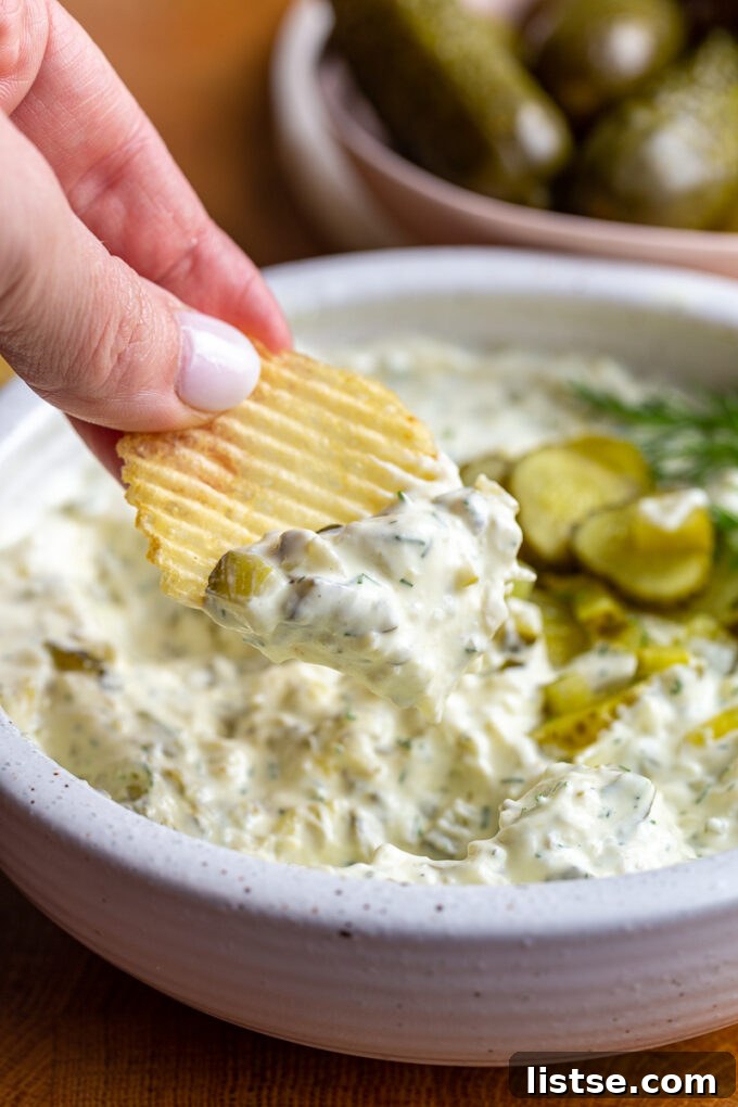 Dill pickle dip in a bowl topped with bright green diced pickles. There's a hand hold a chip up that has some of the dip on it