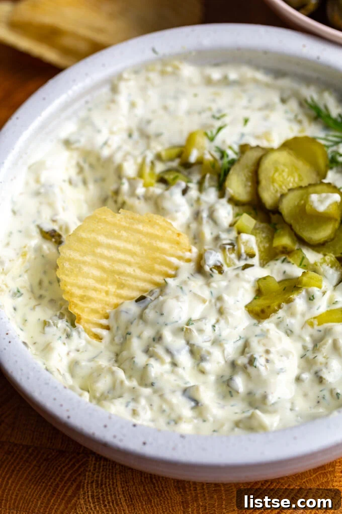 Dill pickle dip in a bowl topped with bright green diced pickles. There is a chip dipped in the bowl