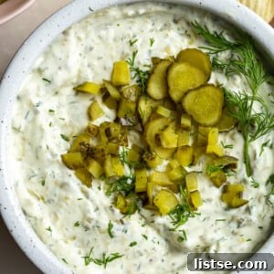 Dill pickle dip in a bowl topped with bright green diced pickles. There are ruffles chips in the corner of the shot