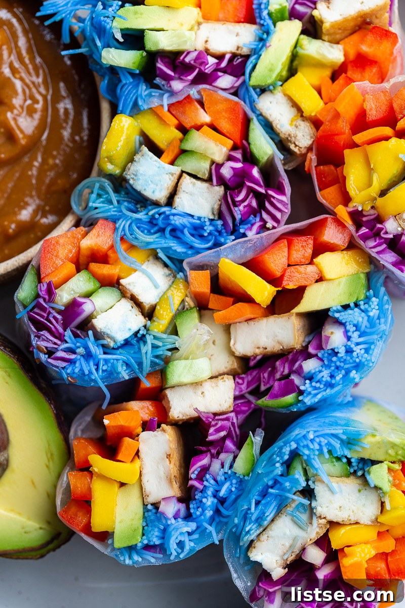 Close-up of freshly made spring rolls, filled with an abundance of colorful vegetables including red bell pepper, carrots, mango, avocado, cucumber, purple cabbage, and distinct blue rice noodles, alongside tofu pieces. The fresh ingredients are clearly visible through the translucent rice paper.