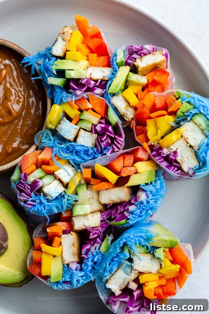A pair of beautifully rolled rainbow spring rolls, showcasing layers of colorful vegetables and blue noodles, with a small dish of creamy peanut sauce and fresh avocado slices in the background. The detailed texture of the rice paper is visible.