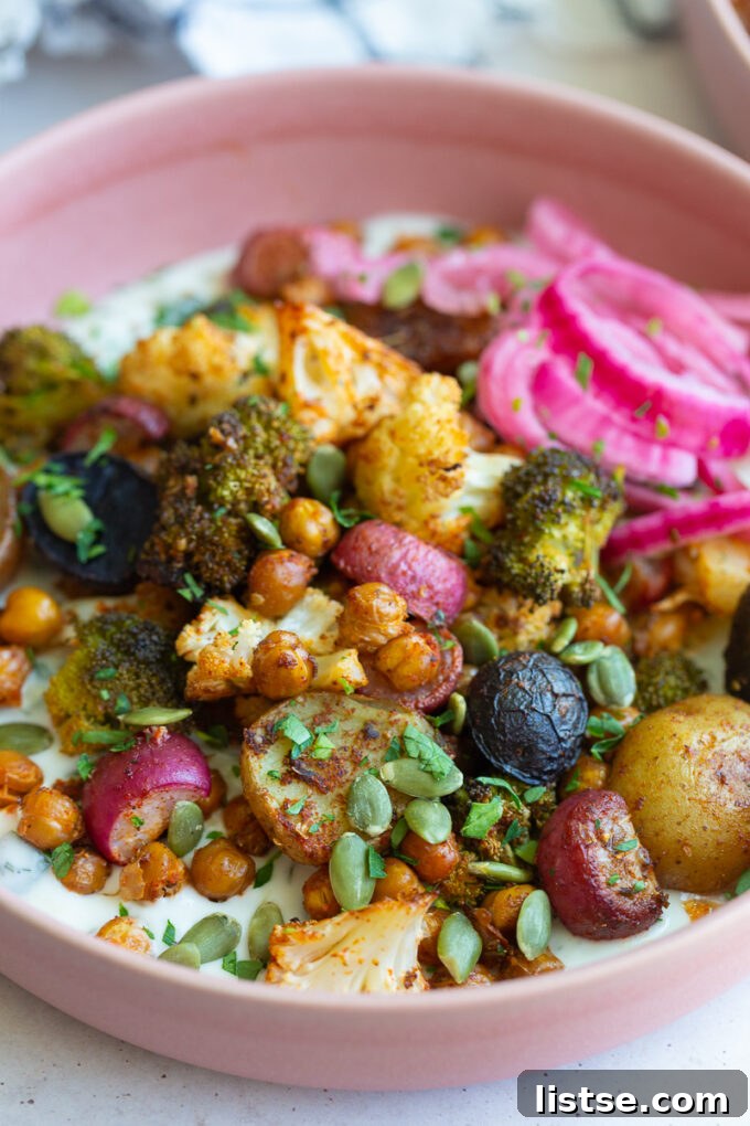 Close-up of a pink bowl filled with an inviting array of roasted vegetables, including golden-brown chickpeas, tender cauliflower, vibrant broccoli, and savory potatoes, all resting on a bed of cool tzatziki with a side of bright pickled red onion.