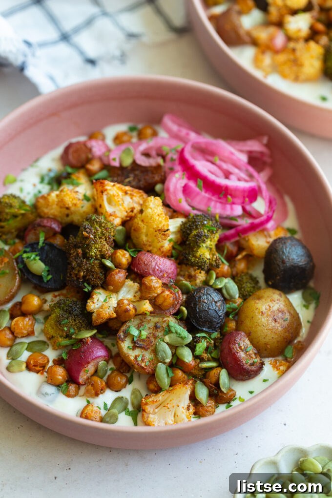 A vibrant pink bowl showcasing the final Roasted Veggie Tzatziki Bowl, with expertly roasted chickpeas, cauliflower, broccoli, and potatoes generously piled over a creamy tzatziki base, garnished with fresh herbs and a side of pickled red onions.