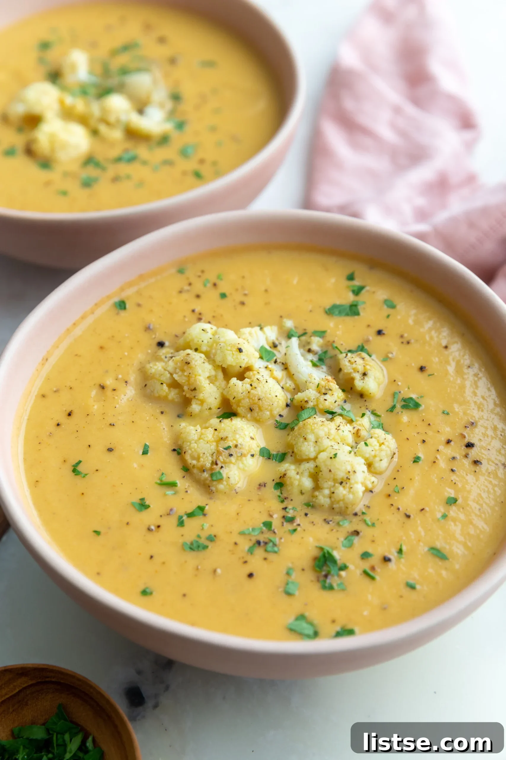 Golden Roasted Cauliflower Velouté 3 Close-up shot of a large pink bowl filled with orange roasted cauliflower soup, adorned with delicate roasted cauliflower pieces and fresh herbs