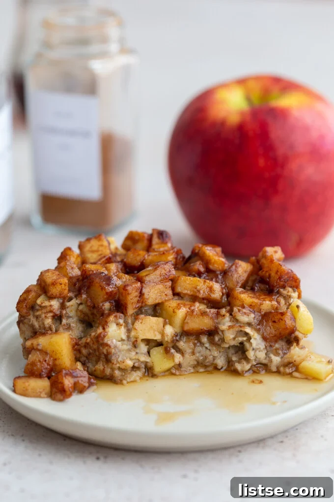 Apple Crumble Baked Oatmeal 3 a close-up of baked oatmeal topped with cinnamon apple chunks, with a portion removed to show the creamy texture inside, and a fresh red apple in the background