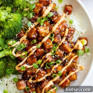 honey garlic tofu in a bowl on top of white rice with cooked broccoli next to it. It's covered in green onion, sesame seeds, and a drizzle of spicy mayo sauce.