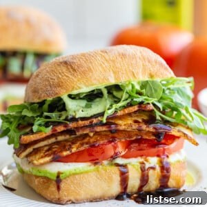 tofu caprese sandwich made with a ciabatta roll and vibrant fresh produce. there's another sandwich and a fresh tomato in the background