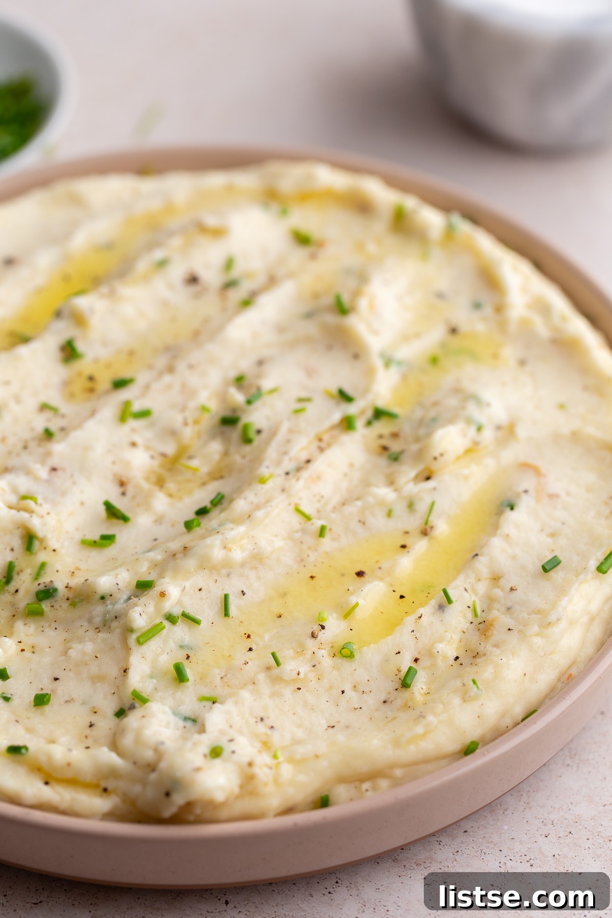 Golden Roasted Garlic Mash 5 A beautifully swirled serving of roasted garlic mashed potatoes in a pink bowl, topped generously with chopped fresh chives, with a small bowl of chives in the corner.
