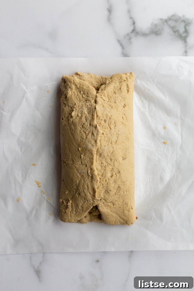 Stuffed Vegan Holiday Roast 6 The second step in creating a seitan roast, illustrating how the seitan dough is carefully folded over the stuffing, partially enclosing it on a piece of parchment paper.