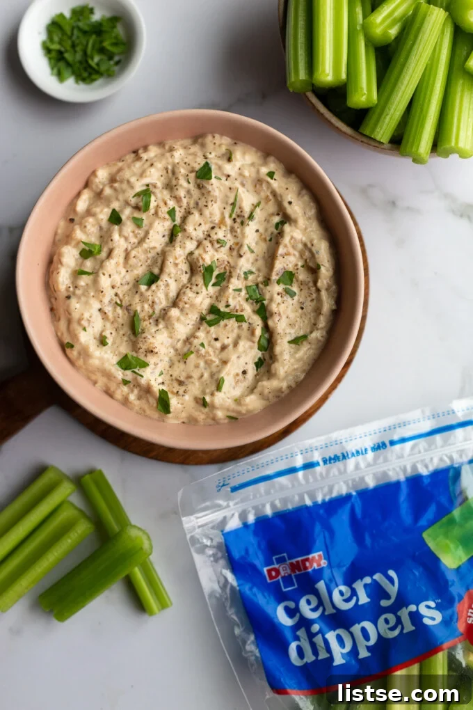 A close-up of a pink bowl filled with creamy French onion dip, surrounded by a generous serving of fresh Dandy celery sticks and a branded bag of celery in the background, all set for a delightful snack.