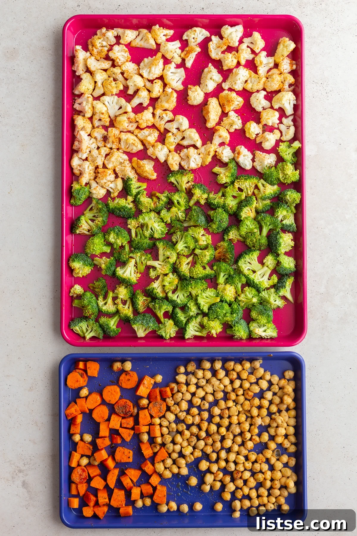 A large red baking sheet filled with seasoned cauliflower and broccoli, with a smaller blue baking sheet below holding carrots and chickpeas, all prepared for roasting