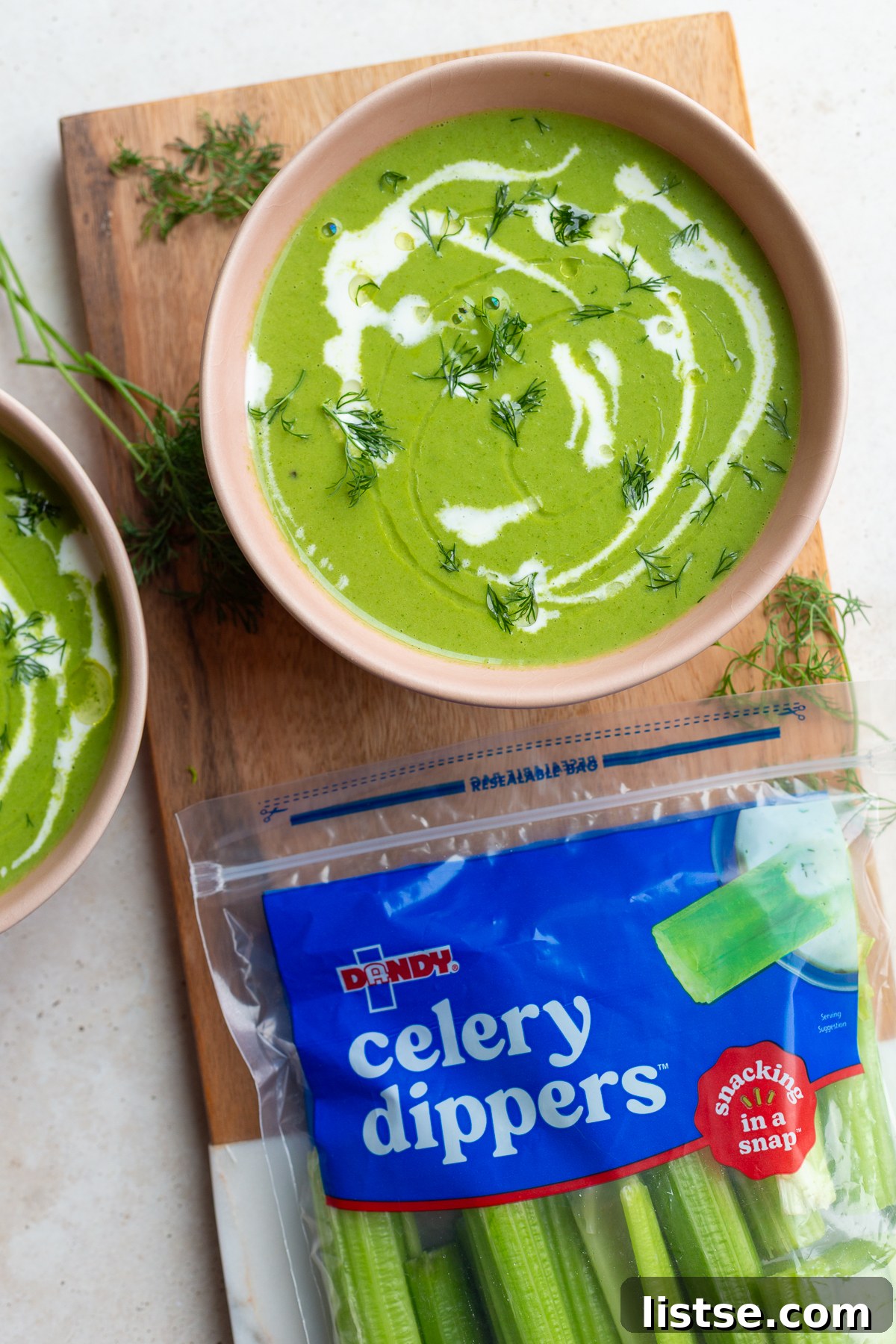 Green Goddess Soup in a pink bowl with a cream drizzle, alongside a bag of Dandy Celery Dippers, highlighting the freshness of the ingredients.