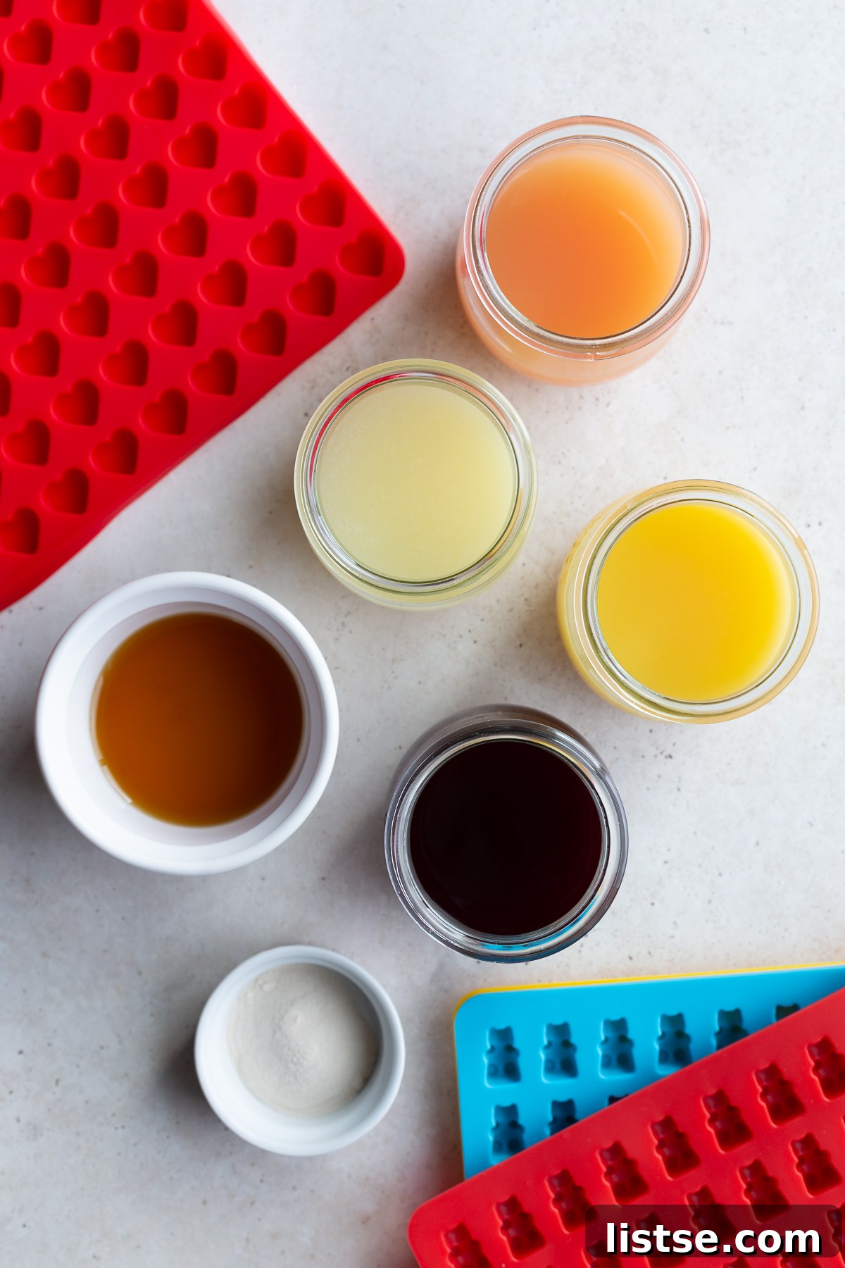 A flat lay photograph featuring small glass bowls containing pure fruit juice, agar-agar powder, and maple syrup, artfully arranged beside various silicone candy molds, highlighting the simplicity of the ingredients for homemade vegan gummies.