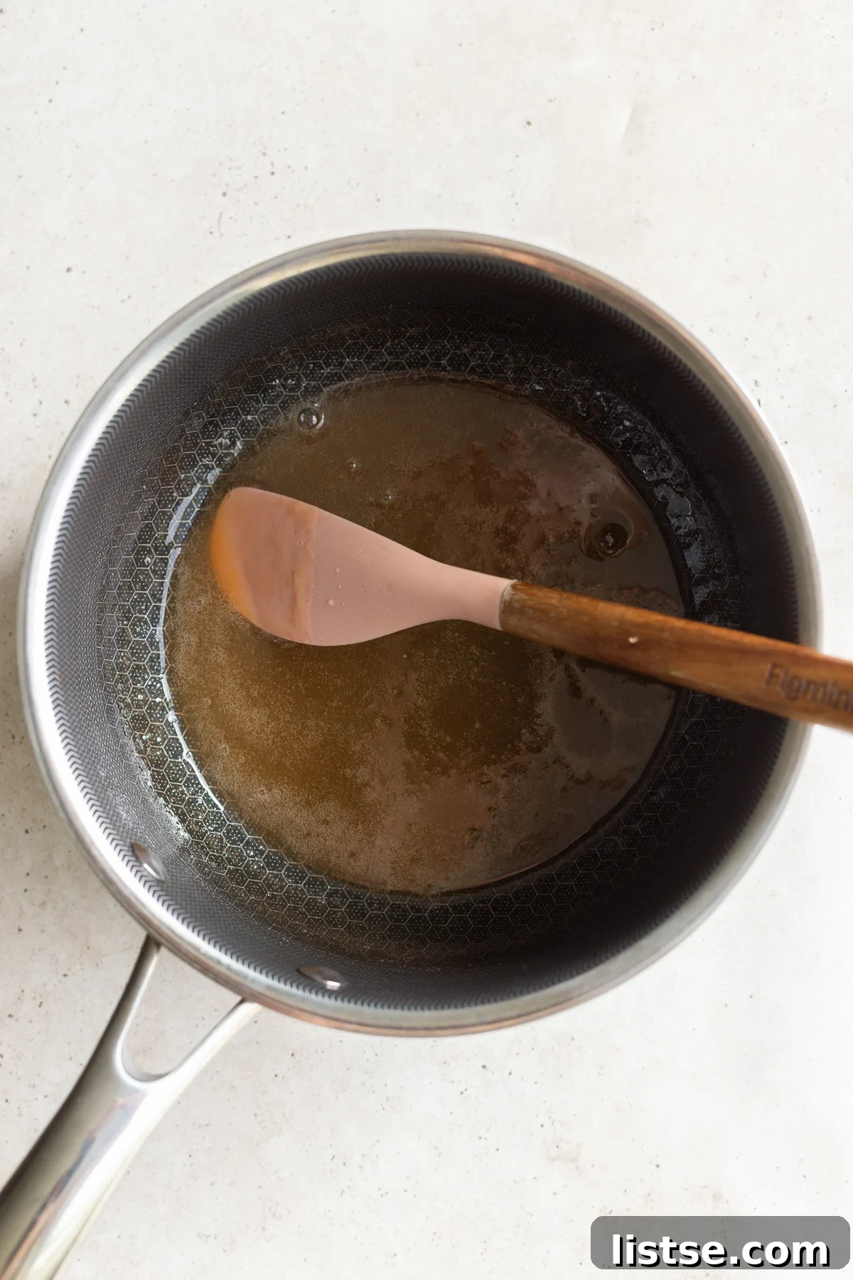 A close-up shot of a stainless steel saucepan on a stovetop, with a whisk gently stirring a vibrant orange liquid mixture, indicating the simmering stage of making vegan gummy bears.