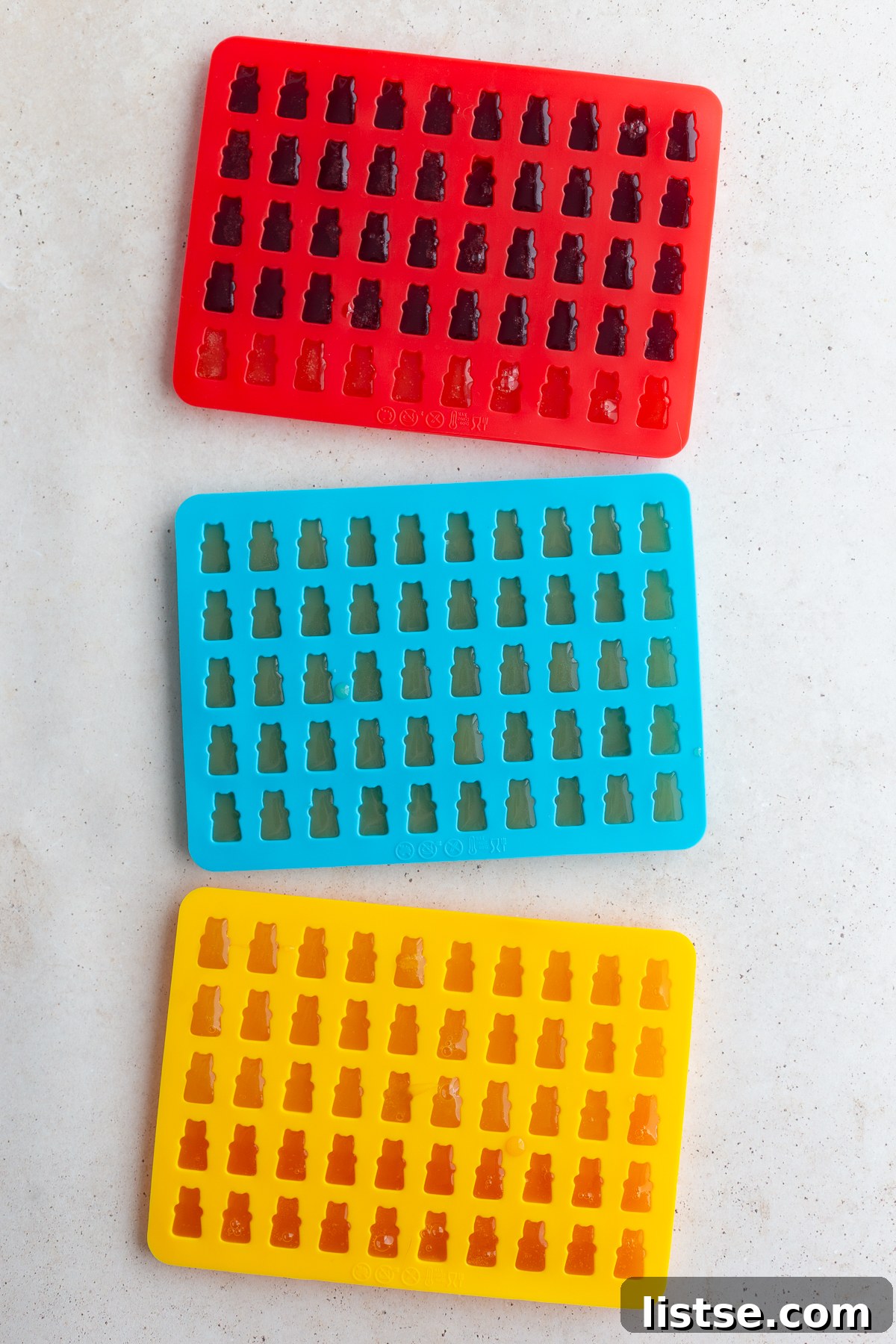 Rows of clear silicone gummy bear molds filled with a bright orange liquid, captured mid-process as the gummy mixture is being transferred into the intricate cavities.