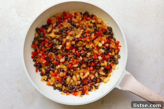 Close-up view of diced onions and red bell peppers expertly sautéing in a large skillet, forming the aromatic base for the vegetarian breakfast burrito filling. Spices are ready to be added for enhanced flavor.