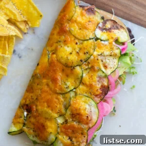 cheesy zucchini wrap stuffed with vegan meat, red onion, avocado and tomato. The wrap is on a plate with chips