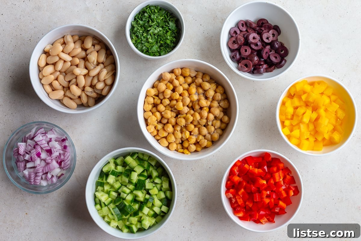 All the fresh salad ingredients laid out: chickpeas, cannellini beans, colorful bell peppers, cucumbers, red onion, fresh parsley, olives, and feta cheese.