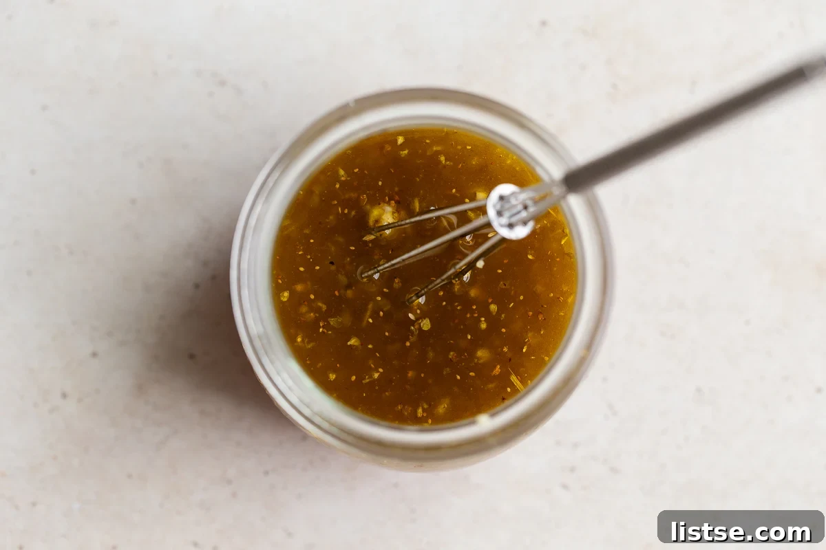 A small jar containing the whisked red wine vinegar, honey, oregano, garlic, salt, and pepper, with olive oil being slowly poured in to create the dressing.