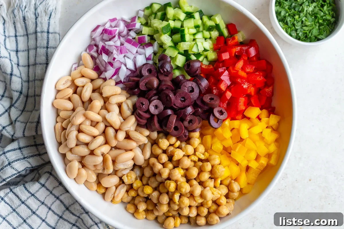 A large mixing bowl filled with the prepared salad ingredients: chickpeas, cannellini beans, colorful bell peppers, cucumbers, red onion, parsley, olives, and crumbled feta cheese.