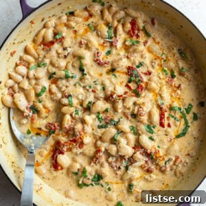 A large skillet brimming with creamy 'Marry Me Butter Beans,' featuring tender butter beans, sun-dried tomatoes, and fresh basil, with a serving spoon nearby.