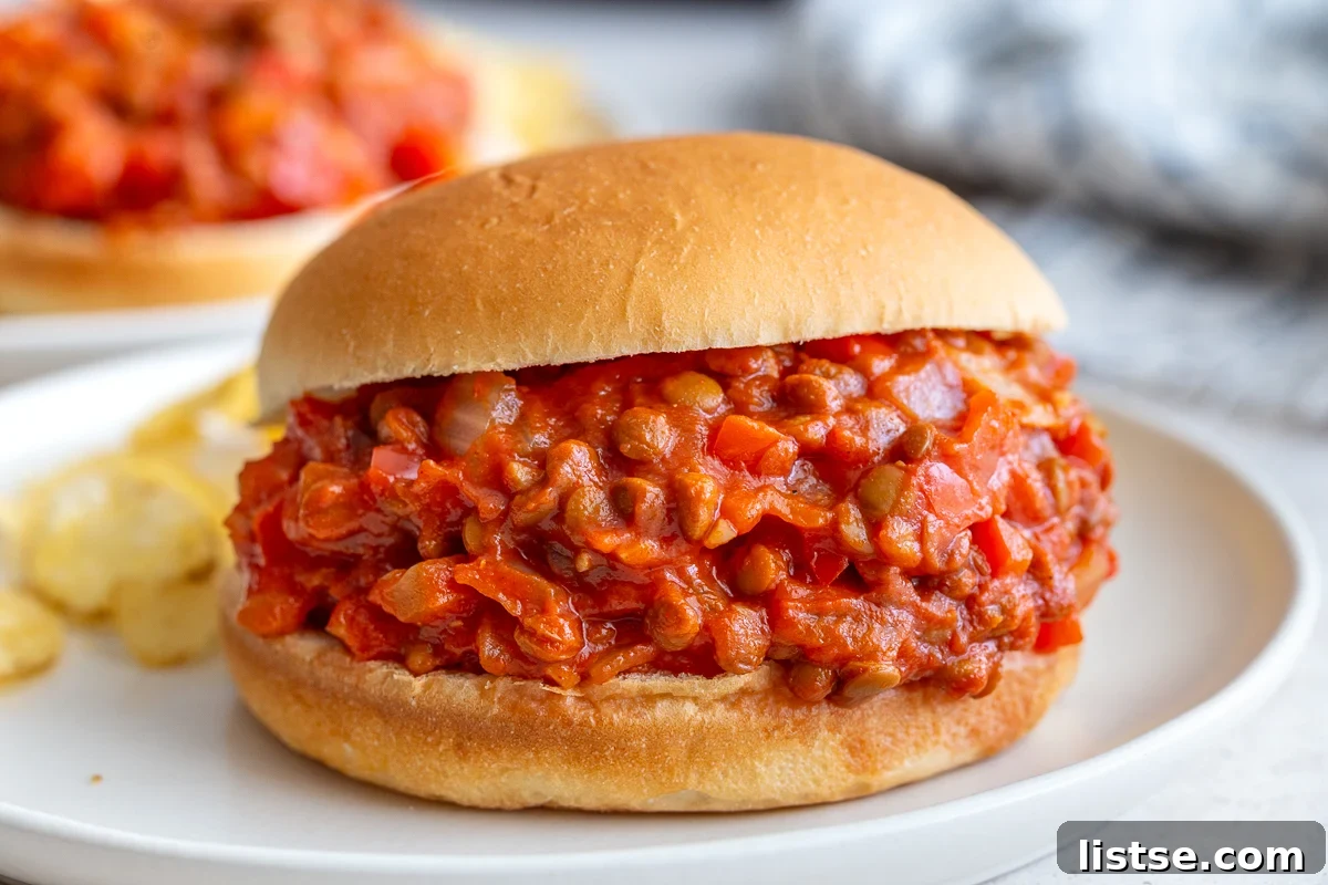 Two lentil sloppy joes on buns, one in focus on a white plate, the other blurred in the background, showcasing the saucy texture.