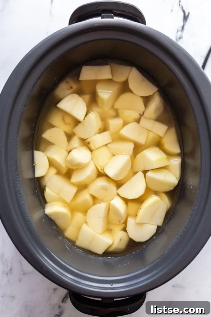 Velvety Slow Cooker Mashed Potatoes 4 Large chunks of peeled russet potatoes simmering in broth and water inside a slow cooker