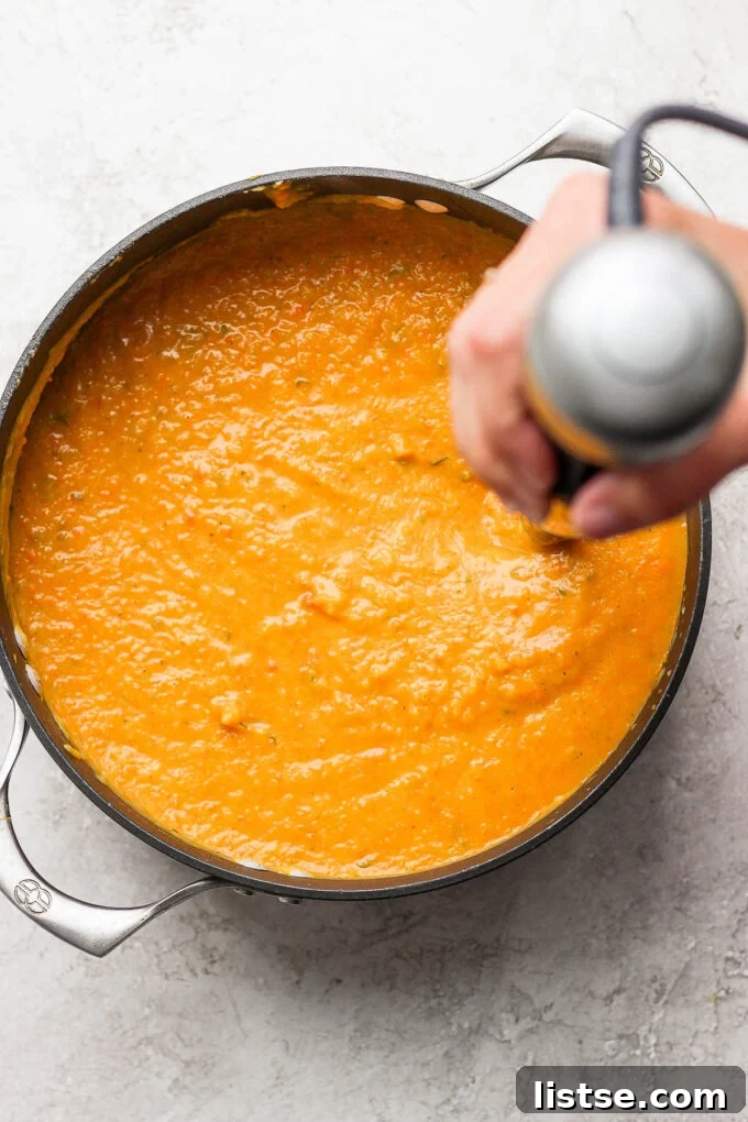 An immersion blender actively pureeing a pot of vibrant orange acorn squash soup until smooth and creamy.