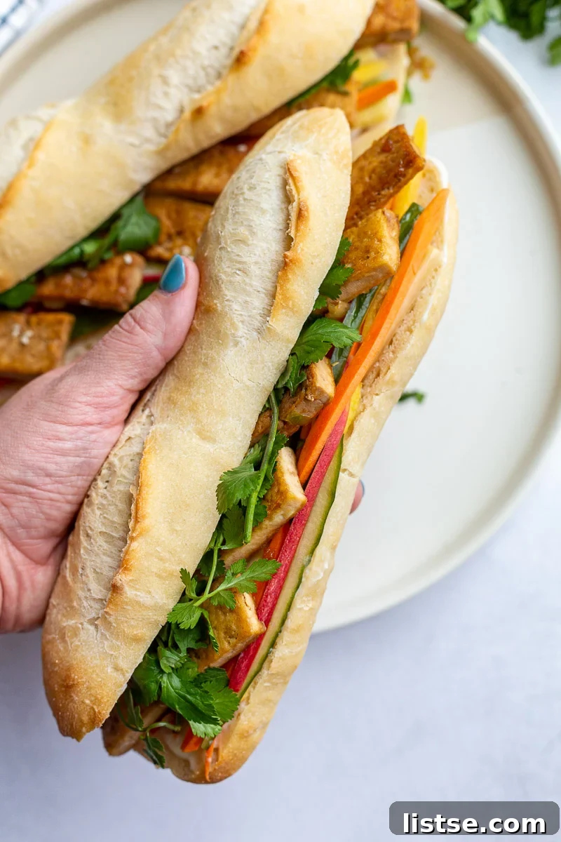 A woman's hand gently holding a freshly made Tofu Banh Mi Sandwich, showcasing the vibrant fillings and fresh cilantro.