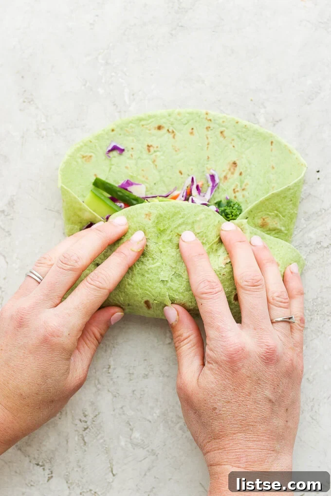 Hands carefully folding a green spinach wrap, tucking in the colorful fresh vegetables and hummus to create a neatly sealed, delicious meal.