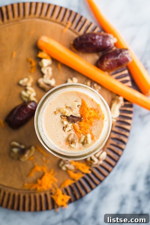 A Carrot Cake Smoothie in a clear glass on a rustic wooden surface, garnished with grated carrots and a cinnamon stick.