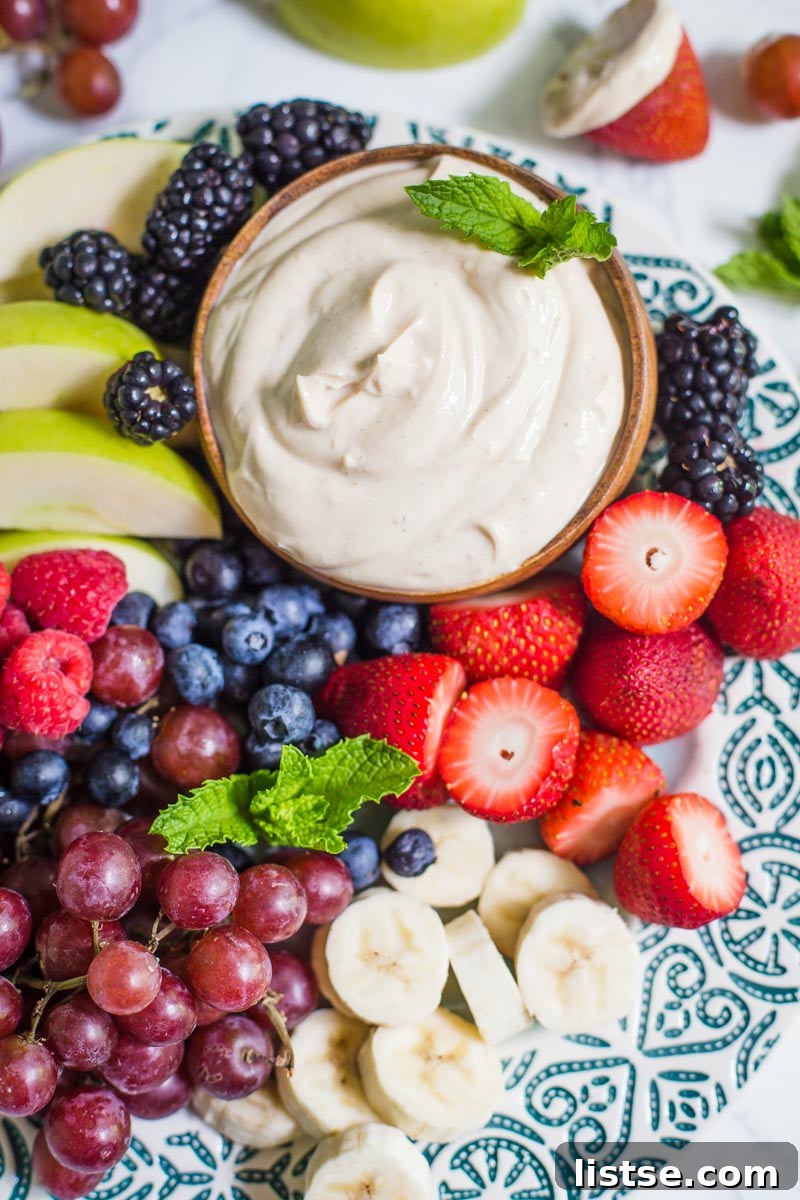 Small bowl of yogurt fruit dip on a platter that's surrounded by colorful fresh fruits including black berries, apples slices, strawberries, and blueberries