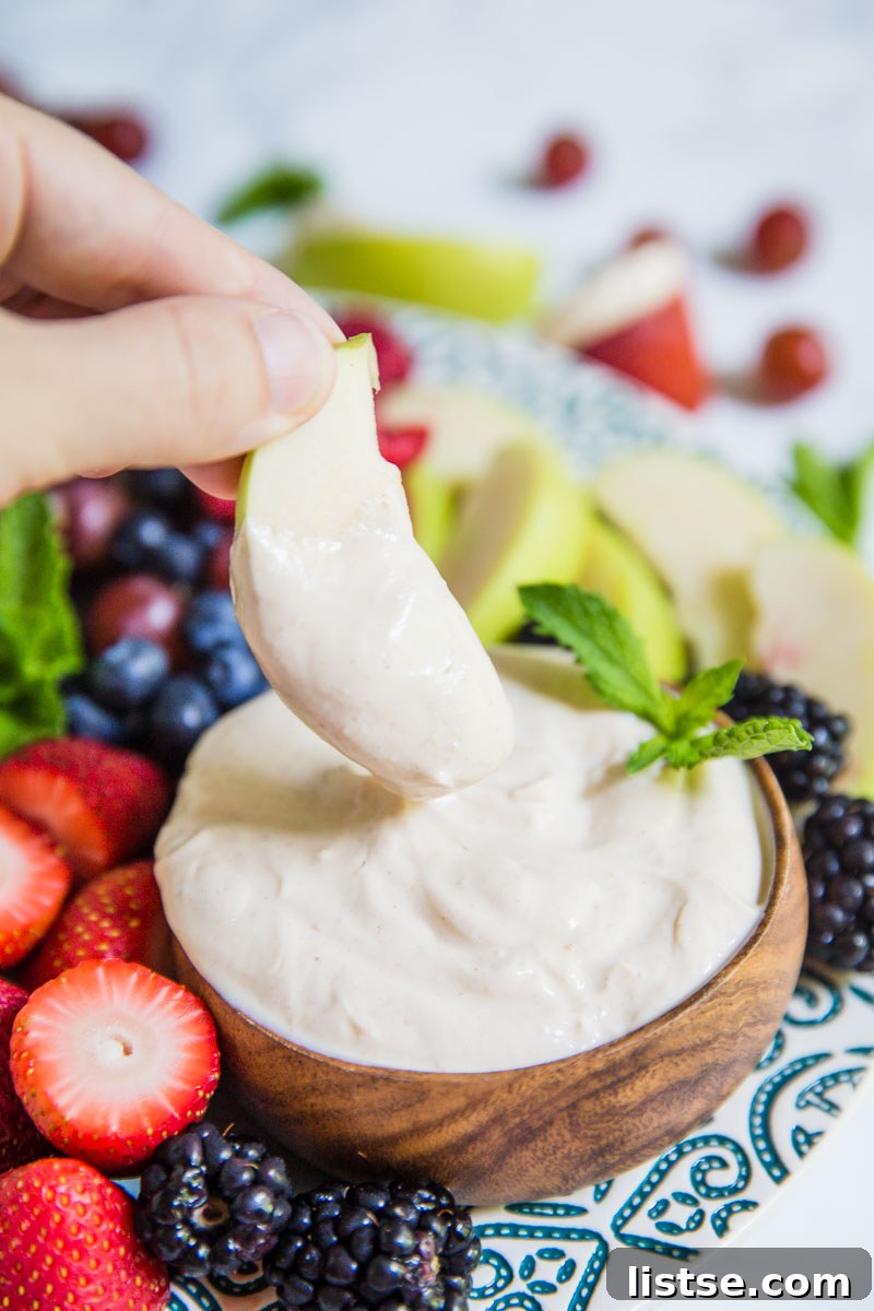Small bowl of yogurt fruit dip on a platter that's surrounded by colorful fresh fruits including black berries, apples slices, strawberries, and blueberries. There's a hand holding an apple slice that has been dipped in the dip