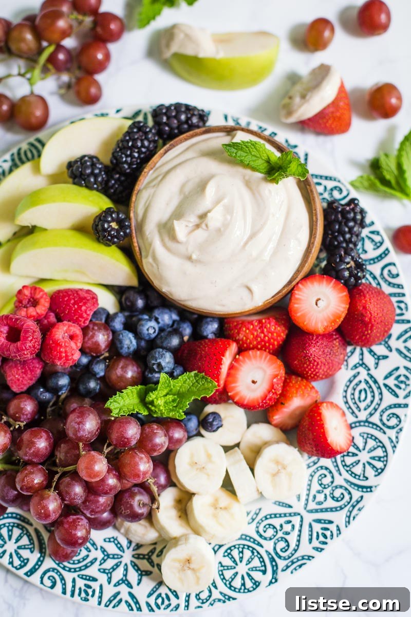 Small bowl of yogurt fruit dip on a platter that's surrounded by colorful fresh fruits including black berries, apples slices, strawberries, and blueberries