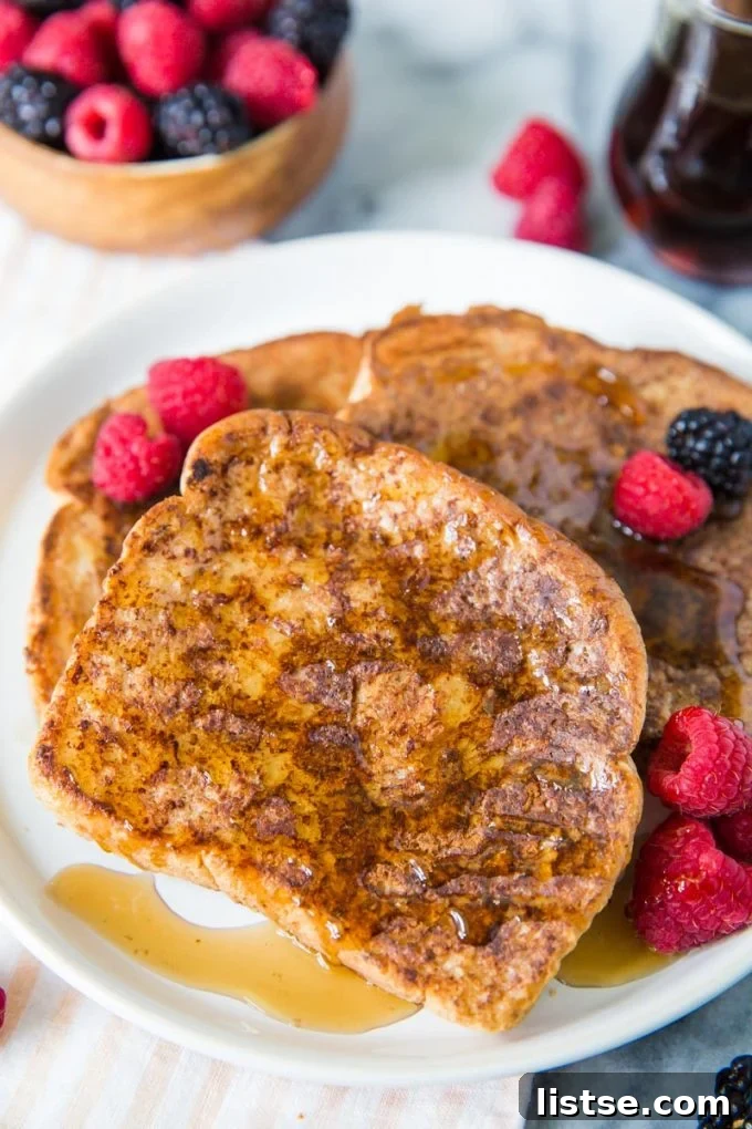 Several golden-brown slices of vegan French toast, generously drizzled with maple syrup and adorned with fresh raspberries and blackberries, arranged on a pristine white plate.