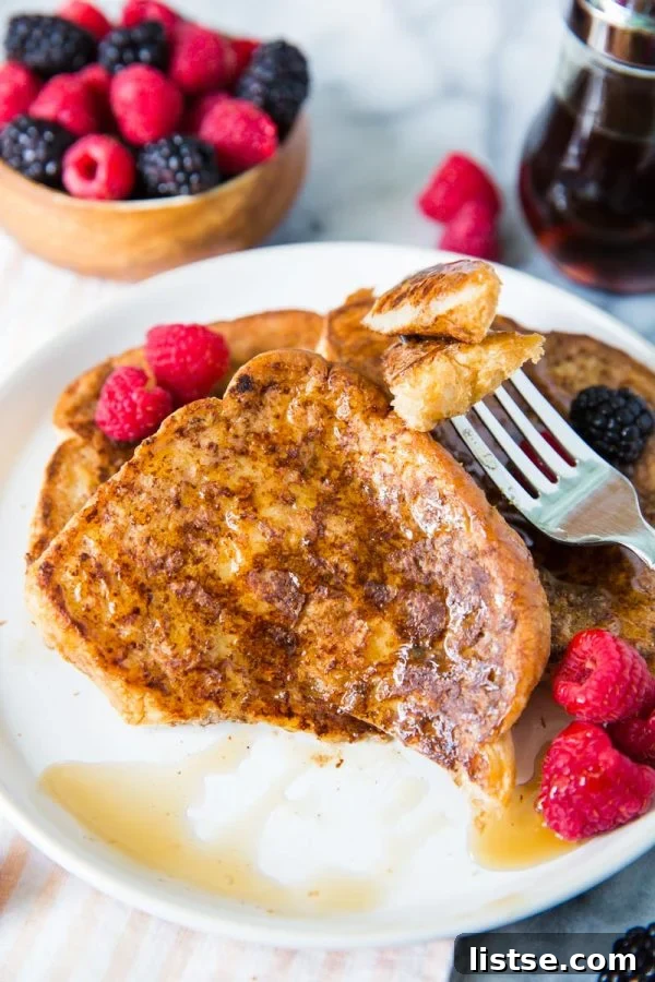Appetizing slices of golden-brown vegan French toast, garnished with fresh raspberries and blackberries and generously coated in maple syrup, served on a white plate with one piece showing a bite taken out of it.