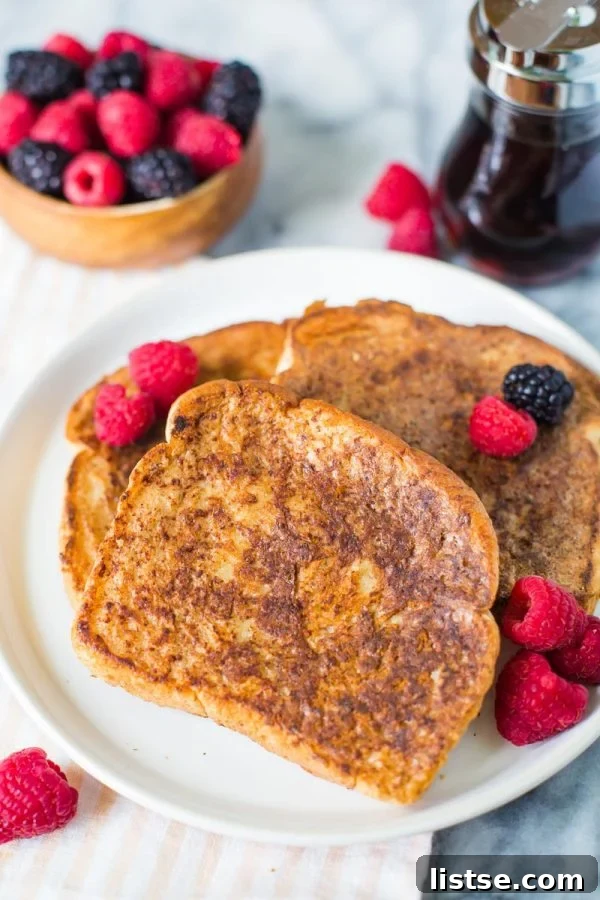 Several perfectly cooked slices of vegan French toast, lightly dusted with powdered sugar and topped with fresh raspberries and blackberries, presented on a clean white plate.